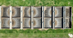 An aerial view of a wastewater treatment facility featuring eight large, rectangular sedimentation tanks arranged in two rows. Each tank contains circulating water, likely part of the treatment process. The tanks are surrounded by green grass, giving a structured and organized appearance.