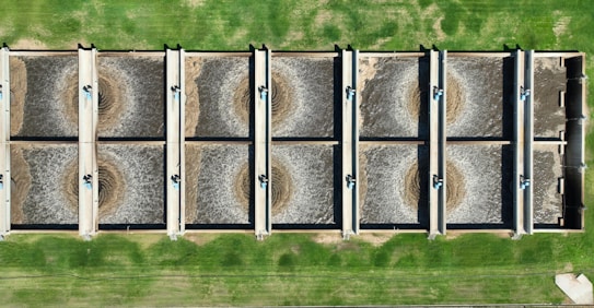 An aerial view of a wastewater treatment facility featuring eight large, rectangular sedimentation tanks arranged in two rows. Each tank contains circulating water, likely part of the treatment process. The tanks are surrounded by green grass, giving a structured and organized appearance.