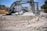 A construction site with a large yellow and gray excavator prominently placed in the foreground. There are piles of rubble and debris scattered around. In the background, several multi-story residential buildings are visible. A few people are seen near the excavator, and a white van is parked nearby.