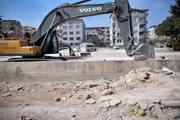 A construction site with a large yellow and gray excavator prominently placed in the foreground. There are piles of rubble and debris scattered around. In the background, several multi-story residential buildings are visible. A few people are seen near the excavator, and a white van is parked nearby.
