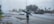 A person wearing a raincoat is standing on a wet road during heavy rain. Cars are visible driving on the road, and there are buildings and trees in the background. The sky appears overcast.