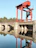 A large red industrial crane is situated on a concrete structure over a body of water, reflecting below. The background consists of a serene landscape with a dense tree line under a clear blue sky.