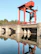 A large red industrial crane is situated on a concrete structure over a body of water, reflecting below. The background consists of a serene landscape with a dense tree line under a clear blue sky.