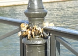 A collection of padlocks attached to the railing of a bridge, symbolizing love or commitment. The locks are affixed to a metal post, overlooking a body of water.