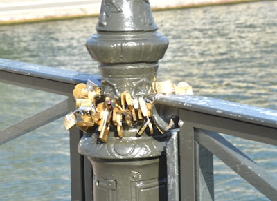 A collection of padlocks attached to the railing of a bridge, symbolizing love or commitment. The locks are affixed to a metal post, overlooking a body of water.