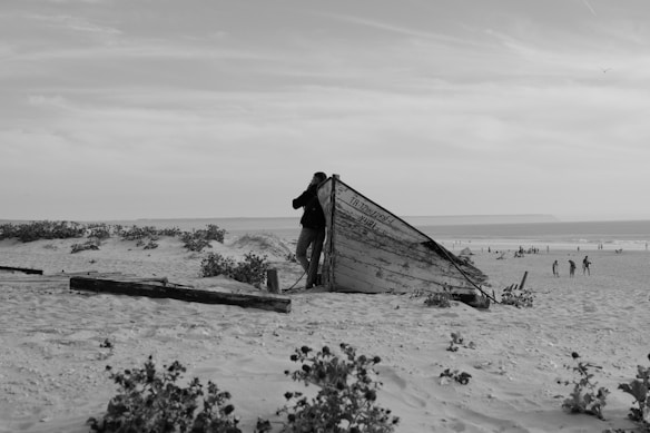 A black and white photograph of a beach scene, featuring an old, weathered boat partially buried in the sand. The boat has text on its side and a person appears to be examining it closely. The beach stretches out to the ocean in the background, where several groups of people are dispersed along the shoreline. Sparse vegetation and clouds are visible in the sky.