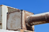 A large, rusted metal duct system against a clear blue sky. The structure includes rectangular and cylindrical sections showing significant signs of rust and weathering. The metal appears aged and worn, adding a rugged, industrial feel to the image.