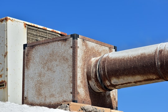 A large, rusted metal duct system against a clear blue sky. The structure includes rectangular and cylindrical sections showing significant signs of rust and weathering. The metal appears aged and worn, adding a rugged, industrial feel to the image.