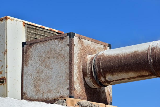 A large, rusted metal duct system against a clear blue sky. The structure includes rectangular and cylindrical sections showing significant signs of rust and weathering. The metal appears aged and worn, adding a rugged, industrial feel to the image.