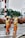Two workers wearing orange safety suits and yellow helmets are on the road. One is holding a broom and appears to be sweeping, while the other holds a wand or baton. They both wear green reflective vests. There are vehicles and green foliage in the background.