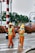 Two workers wearing orange safety suits and yellow helmets are on the road. One is holding a broom and appears to be sweeping, while the other holds a wand or baton. They both wear green reflective vests. There are vehicles and green foliage in the background.
