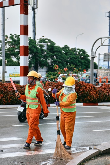 Two workers wearing orange safety suits and yellow helmets are on the road. One is holding a broom and appears to be sweeping, while the other holds a wand or baton. They both wear green reflective vests. There are vehicles and green foliage in the background.