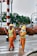 Two workers wearing orange safety suits and yellow helmets are on the road. One is holding a broom and appears to be sweeping, while the other holds a wand or baton. They both wear green reflective vests. There are vehicles and green foliage in the background.