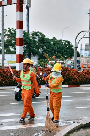 Two workers wearing orange safety suits and yellow helmets are on the road. One is holding a broom and appears to be sweeping, while the other holds a wand or baton. They both wear green reflective vests. There are vehicles and green foliage in the background.