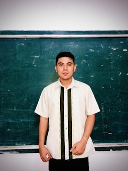 A person stands in front of a dark green chalkboard, wearing a light-colored short-sleeve shirt with dark vertical stripes. The chalkboard has visible chalk marks and smudges.