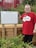 A person wearing a red shirt with 'Bay de Verde Heritage Premises' stands beside a memorial plaque for Glen Roy Blundon. The plaque describes Glen's life, his challenges with muscular dystrophy, and his accomplishments. The setting includes a wooden structure and green foliage, with a red structure in the background.