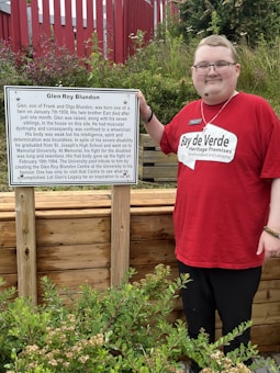 A person wearing a red shirt with 'Bay de Verde Heritage Premises' stands beside a memorial plaque for Glen Roy Blundon. The plaque describes Glen's life, his challenges with muscular dystrophy, and his accomplishments. The setting includes a wooden structure and green foliage, with a red structure in the background.