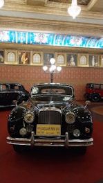 A classic Jaguar MK VIII car is displayed in an opulently decorated room with a high ceiling adorned with chandeliers. The room has ornate wall art featuring portraits, and the floor is covered with a deep red carpet. The black Jaguar, with gleaming chrome details, is prominently positioned, contrasting with the richly patterned blue and gold ceiling decorations.
