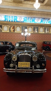 A classic Jaguar MK VIII car is displayed in an opulently decorated room with a high ceiling adorned with chandeliers. The room has ornate wall art featuring portraits, and the floor is covered with a deep red carpet. The black Jaguar, with gleaming chrome details, is prominently positioned, contrasting with the richly patterned blue and gold ceiling decorations.