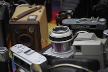 A collection of vintage cameras with various designs and sizes. Prominent models include a dual-lens box camera and a silver camera with a prominent lens. Some cameras have unique features like knobs and dials.
