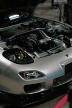 A close-up view of a car engine with the hood open. The car has a sleek silver exterior with distinctive headlight designs. Various engine components, including metal pipes and hoses, are visible, showcasing the complex machinery.
