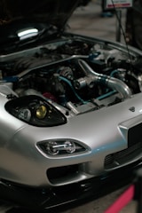 A close-up view of a car engine with the hood open. The car has a sleek silver exterior with distinctive headlight designs. Various engine components, including metal pipes and hoses, are visible, showcasing the complex machinery.