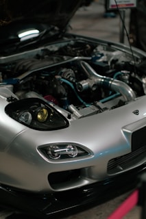 A close-up view of a car engine with the hood open. The car has a sleek silver exterior with distinctive headlight designs. Various engine components, including metal pipes and hoses, are visible, showcasing the complex machinery.
