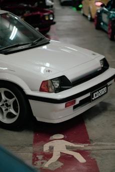 A white vintage car with a black trim and license plate reading JCD 2020 is parked indoors on a concrete floor. The car is alongside other colorful vehicles in a dimly lit garage, and there is a pedestrian symbol painted on the floor.
