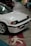 A white vintage car with a black trim and license plate reading JCD 2020 is parked indoors on a concrete floor. The car is alongside other colorful vehicles in a dimly lit garage, and there is a pedestrian symbol painted on the floor.