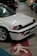 A white vintage car with a black trim and license plate reading JCD 2020 is parked indoors on a concrete floor. The car is alongside other colorful vehicles in a dimly lit garage, and there is a pedestrian symbol painted on the floor.