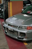 A silver car is parked indoors with a visible license plate reading 'BMM 737'. The vehicle's front grille and hood vents are prominently displayed, and the environment suggests an indoor showroom or parking area with colorful walls in the background.