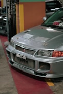 A silver car is parked indoors with a visible license plate reading 'BMM 737'. The vehicle's front grille and hood vents are prominently displayed, and the environment suggests an indoor showroom or parking area with colorful walls in the background.