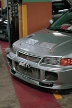 A silver car is parked indoors with a visible license plate reading 'BMM 737'. The vehicle's front grille and hood vents are prominently displayed, and the environment suggests an indoor showroom or parking area with colorful walls in the background.
