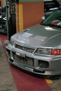 A silver car is parked indoors with a visible license plate reading 'BMM 737'. The vehicle's front grille and hood vents are prominently displayed, and the environment suggests an indoor showroom or parking area with colorful walls in the background.