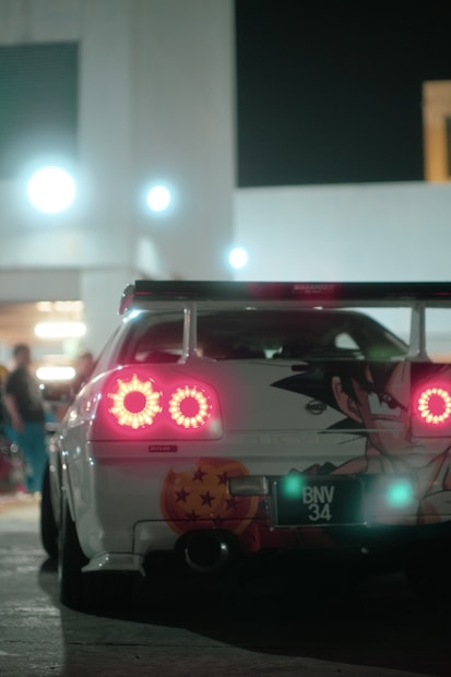 A sports car with vibrant rear lights features an anime character and a Dragon Ball design on the rear. The setting is urban at night with strong artificial lighting, adding to a sleek and modern atmosphere.