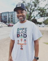 A man is smiling outdoors, wearing a black cap with a graphic on it and a light gray t-shirt that reads 'Dope Black Dad' with colorful text and illustrations of three cartoon characters below, each with different names.