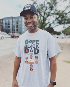 A man is smiling outdoors, wearing a black cap with a graphic on it and a light gray t-shirt that reads 'Dope Black Dad' with colorful text and illustrations of three cartoon characters below, each with different names.