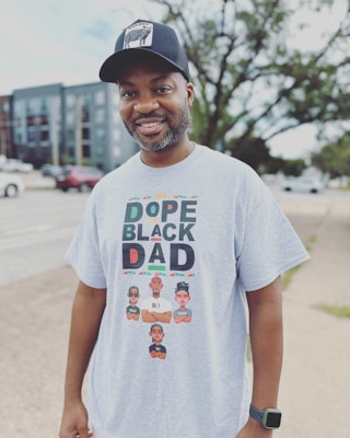 A man is smiling outdoors, wearing a black cap with a graphic on it and a light gray t-shirt that reads 'Dope Black Dad' with colorful text and illustrations of three cartoon characters below, each with different names.