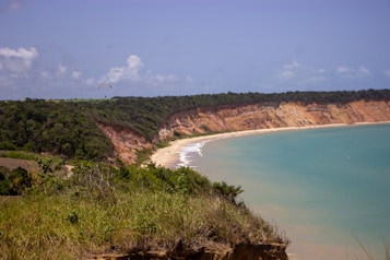 A coastal landscape featuring a high cliff with green vegetation overlooking a serene beach. The cliffs display layers of red and orange hues, while the turquoise ocean gently washes over the sandy shore. In the distance, a paraglider is visible in the sky, adding a touch of adventure to the peaceful scene.