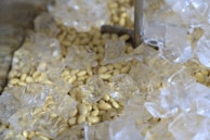 A close-up image of ice cubes scattered over a bed of small, pale yellow beans or seeds. The ice cubes are transparent and glistening, reflecting light and creating a cool atmosphere.