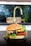 A close-up of a gourmet cheeseburger placed on a dark surface. The burger consists of a whole grain seeded bun, melted cheese, a beef patty, fresh tomato slices, and leafy greens. The background shows a slightly blurred kitchen faucet and sink area.
