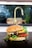A close-up of a gourmet cheeseburger placed on a dark surface. The burger consists of a whole grain seeded bun, melted cheese, a beef patty, fresh tomato slices, and leafy greens. The background shows a slightly blurred kitchen faucet and sink area.