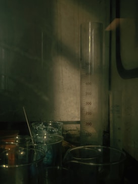 A dimly lit laboratory scene featuring various glassware, including a prominent graduated cylinder with measurement markings. Several smaller beakers and flasks are arranged on a surface, some containing stirring rods. The overall environment appears industrial or scientific with a moody ambiance.
