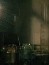 A dimly lit laboratory or scientific setting featuring various glassware and beakers. A tall graduated cylinder partially filled with liquid can be seen in the background, with reflections and shadows creating a moody ambiance.