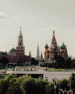 A scenic view of iconic Russian architecture featuring the colorful domes and spires of Saint Basil's Cathedral alongside the red brick towers of the Kremlin. The scene includes lush greenery in the foreground and a bridge with people walking on it.