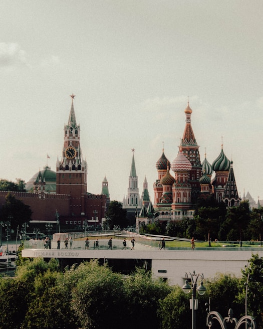 A scenic view of iconic Russian architecture featuring the colorful domes and spires of Saint Basil's Cathedral alongside the red brick towers of the Kremlin. The scene includes lush greenery in the foreground and a bridge with people walking on it.