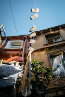 Several pairs of shoes hanging by their laces from wires strung between buildings, with laundry hanging on clotheslines nearby. The buildings feature weathered facades and windows, with a clear blue sky visible above. Green foliage adds a touch of nature to the urban setting.