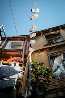 Several pairs of shoes hanging by their laces from wires strung between buildings, with laundry hanging on clotheslines nearby. The buildings feature weathered facades and windows, with a clear blue sky visible above. Green foliage adds a touch of nature to the urban setting.