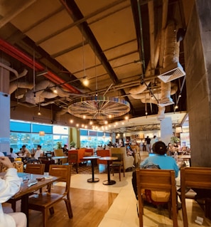 A cozy and modern cafe interior with a high ceiling and exposed ductwork. Warm lighting is cast by multiple hanging lights, and several people are seated at wooden tables. Large windows on one side allow natural light to filter in, creating a welcoming atmosphere.