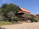 A traditional wooden building with a red-tiled roof, surrounded by lush greenery and trees. The architecture features open spaces and ornate design elements typical of Southeast Asian style. The area is peaceful, with a paved path in the foreground leading up to the structure.